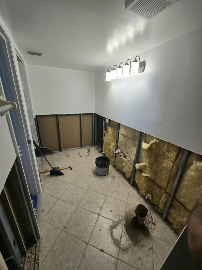 Bathroom wall opened along the bottom after a leak, insulation exposed and plastic sheeting protecting the room